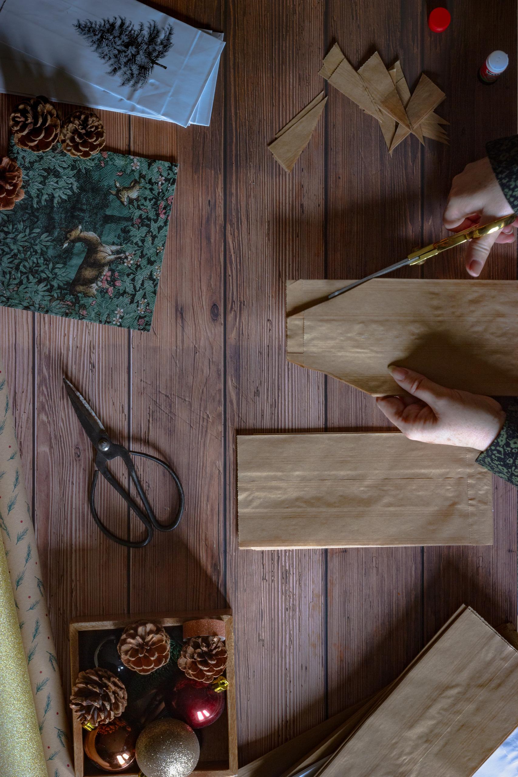 A person cuts parts off brown paper bags. The table has other Christmassy arts and crafts materials on it.