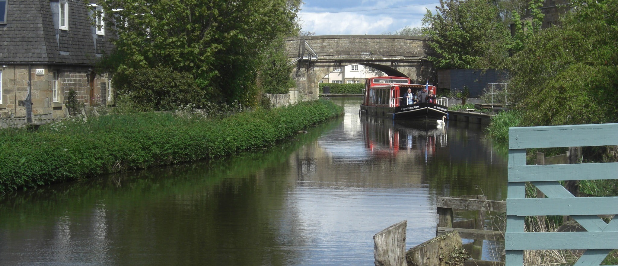 Red barge, with a few people standing at the front, travels slowly down a canal surrounded by green trees.