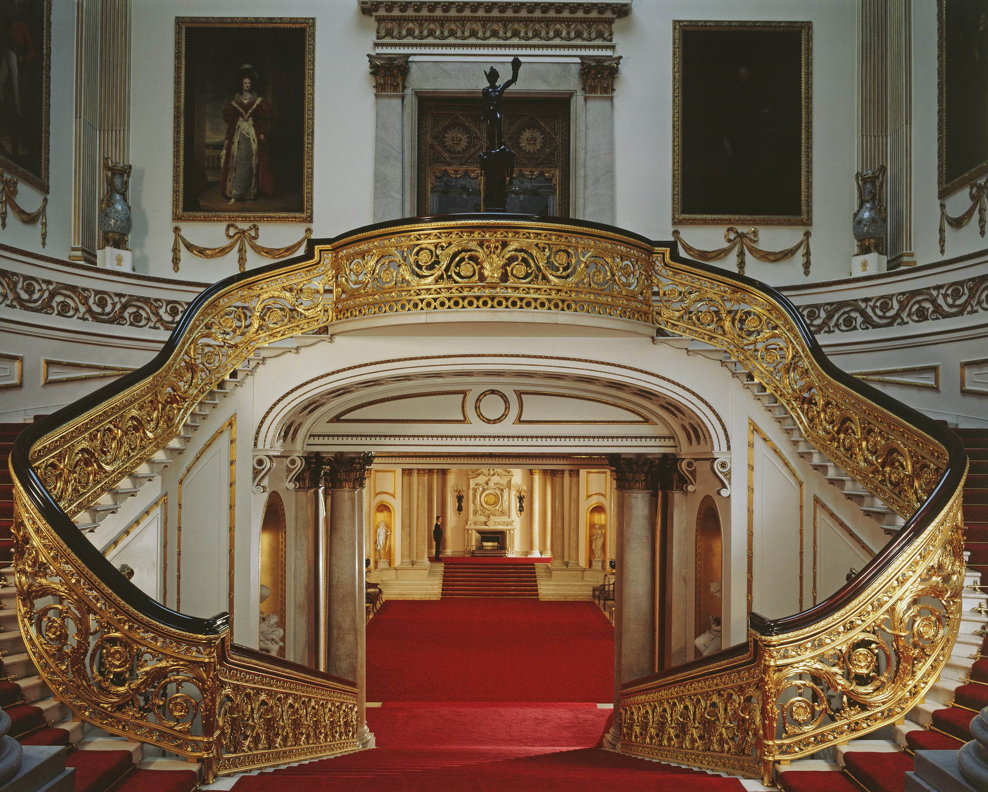Grand staircase in Buckingham Palace, with a red carpet going down the corridor, golden bannisters and statues.