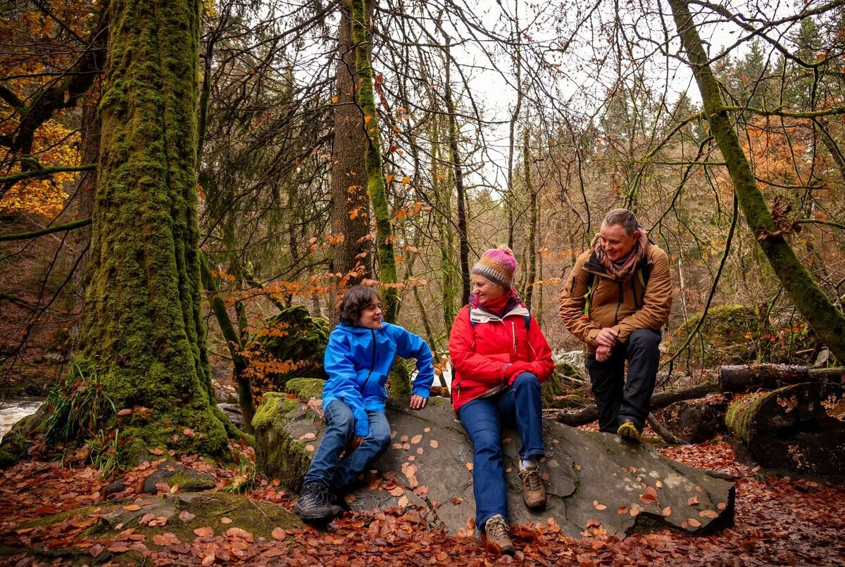 Two adults and a child sit chatting in an autumnal forest.