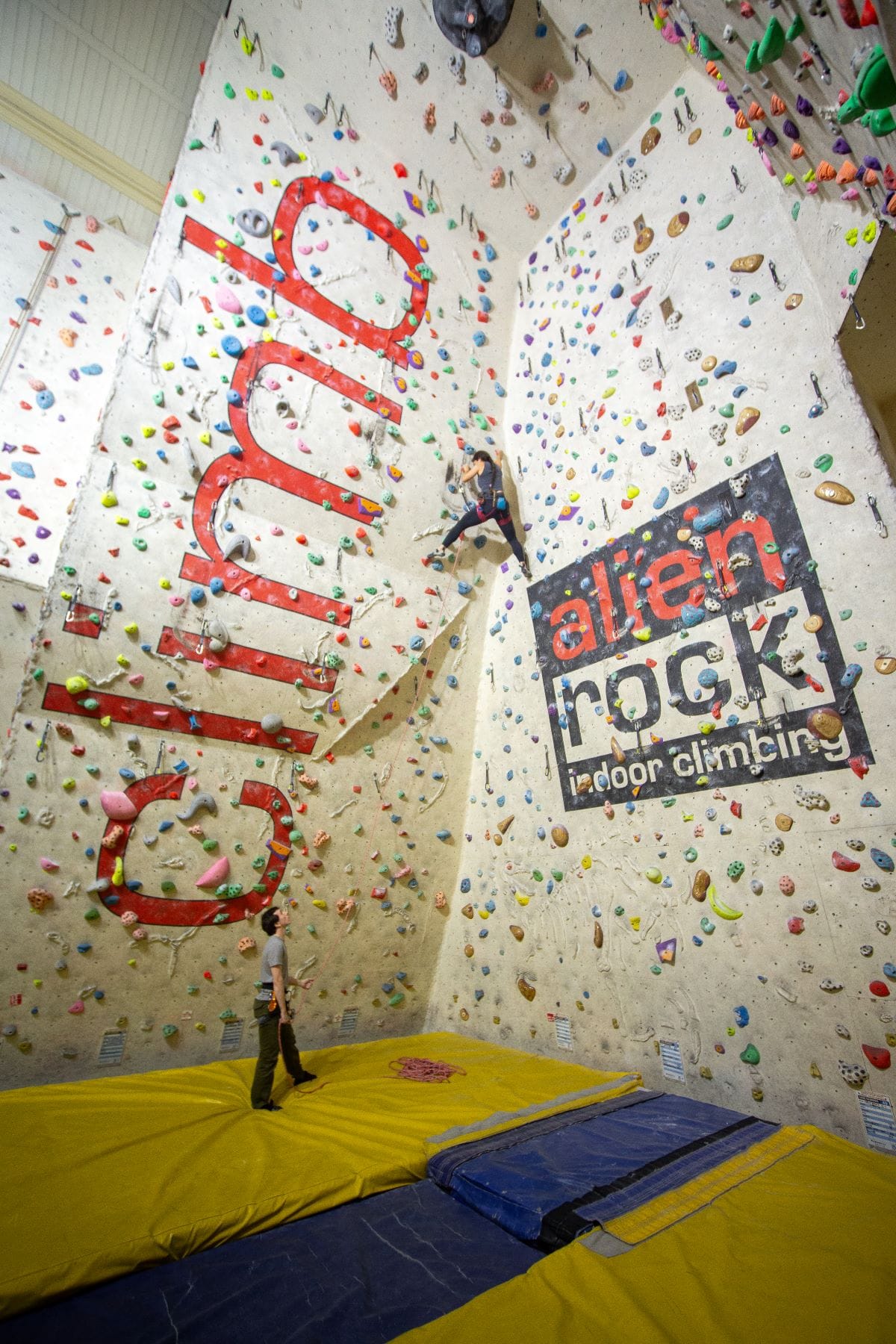 View upwards from the bottom of an indoor climbing wall, which says "alien rock". Someone is halfway up the wall, with someone else controlling the rope from the bottom.