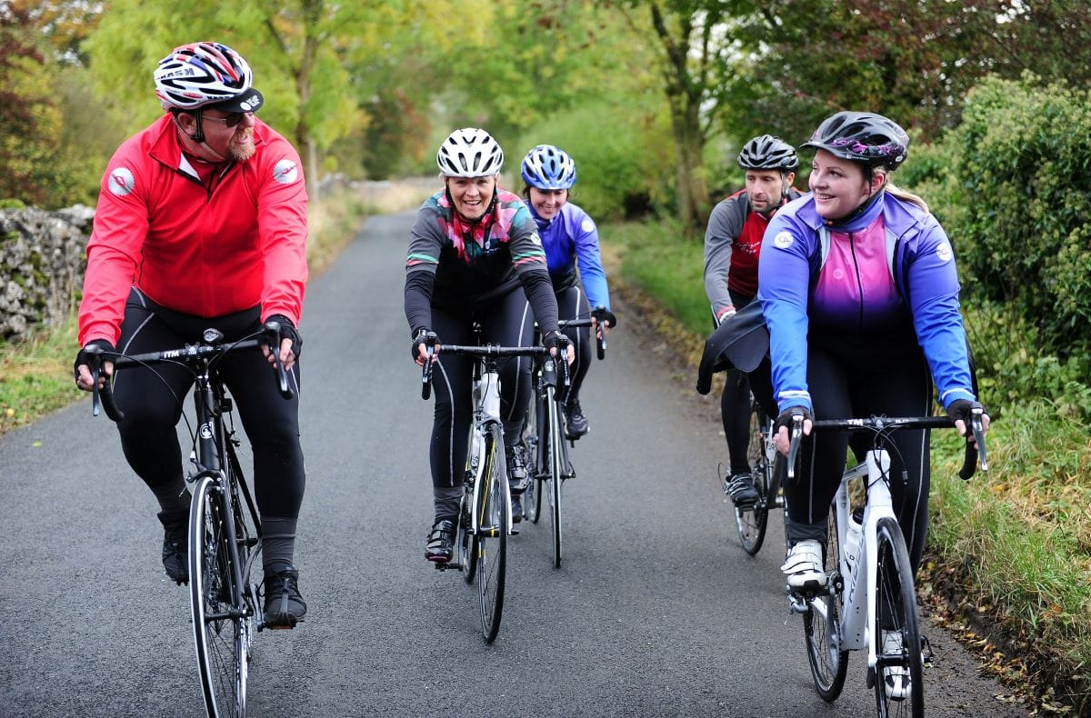 A group of 5 adults on a bike ride, along a road through a wooded area. They are all wearing cycling jerseys and helmets, and chatting together.